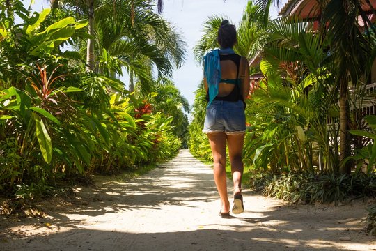 Young girl in shorts walking towards the beach through a sand path surrounded by nature in the island of Koh Phangan, Thailand
