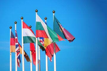 Flags of different countries flutters in the wind against a bright blue sky with copy space