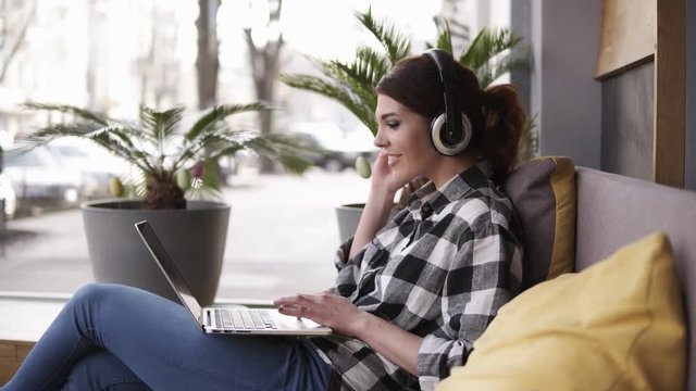 Beautiful, cheerful woman listening to music on laptop at trendy room with headphones sitting on a couch in casual. Happy, enjoy her time. Side view
