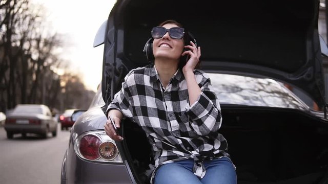 Smiling brunette listen to the music in headphone while sitting in the open car's trunk among the road. Cell phone. Good mood