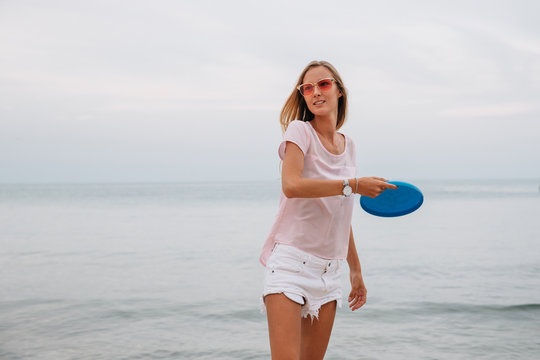 Young charming woman playing frisbee near the sea, holding frisbee disk. Dressed in shorts and t-shirt, in sunglasses.