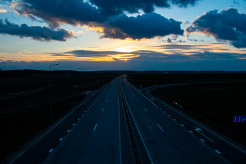 Asphalt road with four lanes leaving the sunset, blue sunset sky with the setting sun, hidden behind the clouds. Clouds illuminated by sunset light
