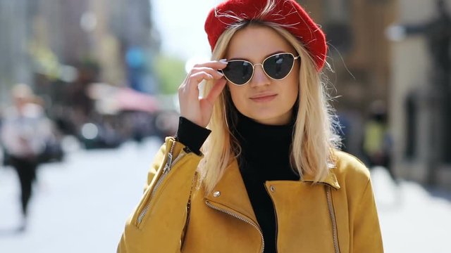 beautiful blonde pretty woman taking off sunglasses wear red beret hat and leather jacket standing outside thoughtful looking at camera attractive female fashionable style cute lady sunlight sunny day