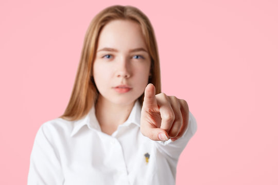 Lovely serious confident young female points at camera with fore figer, chooses someone, isolated on pink background, focus on hand, blurred background. I choose you! People and choice concept