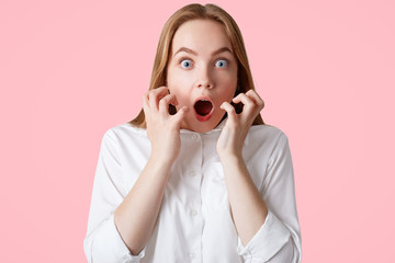 Scared terrified attractive young female looks with wide opened mouth and blue eyes, being frightened of something, gestures actively, wears elegant white shirt, isolated over pink background