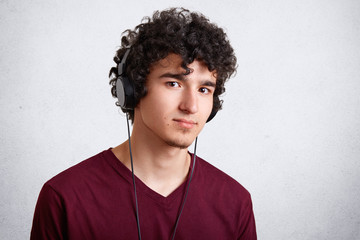 Fototapeta na wymiar Studio shot of male teenager listens music in modern headphones, looks confidently and seriously at camera, poses against white concrete wall, dressed casually. People, youth, leisure concept