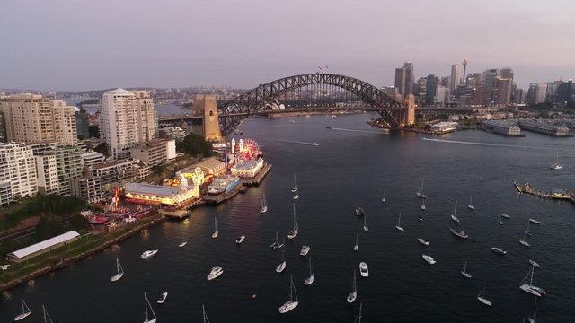 Lavender bay part of Sydney harbour at sunset from arch of Sydney Harbour bridge and distant city CBD towers flying backwards over yachts.
