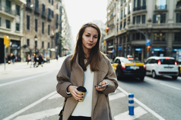 Fototapeta premium Young fashion blogger with long blonde hair is messaging to friend by a smartphone while crossing the road with a paper coffee cup in her hand. Attractive stylish girl is reading sms on mobile phone.