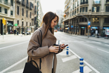 Model look woman reading emails on the smartphone while standing on a street background. Caucasian female texting messages on the mobile phone while crossing the street holding a take away coffee cup.