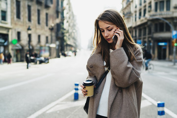 Beautiful woman with long blonde hair calling to the friend by a smartphone while crossing the road with a blank paper cup in her hand. Attractive hipster girl is talking by a mobile phone.