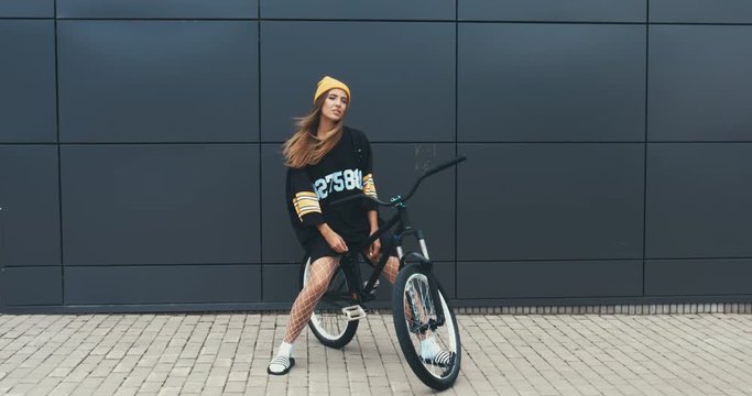 Young stylish Caucasian female sitting on a bicycle against grey wall background. 4K UHD RAW edited footage