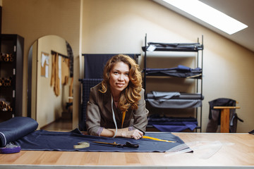 Portrait of professional adult dressmaker female looking at camera, working with a cutting material at tailors workshop