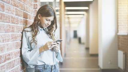 Young girl wearing a white shirt and jeans is looking at her smartphone screen standing in a business building corridor.