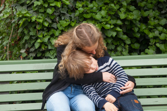 Girl hugs her older brother while they doze sitting on a bench outdoors