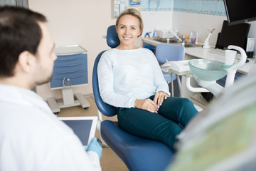 Obraz premium Beautiful woman sitting in chair of dentist smiling at camera while doctor using tablet in cabinet. 