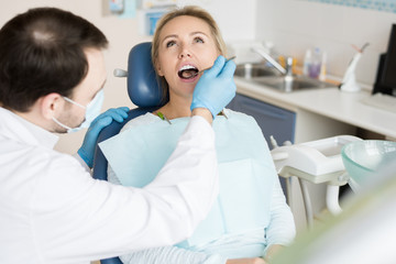 Fototapeta premium Confident man in gloves and mask using small mirror tool and exploring state of teeth of young woman on seat. 