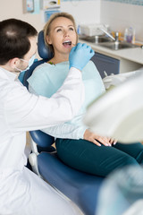 Doctor in uniform and gloves using small mirror tool and checking teeth of young woman on chair. 