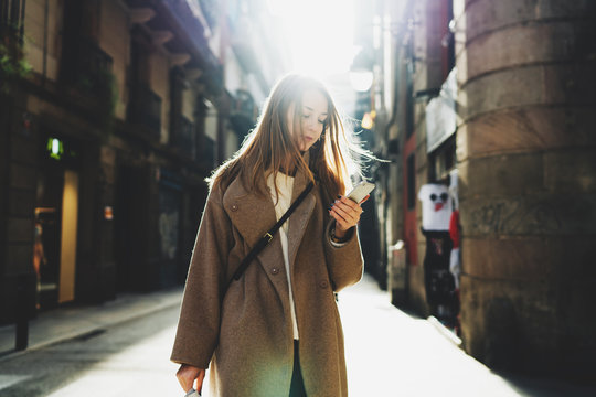 Portrait of young hipster girl holding a mobile phone in her hand looking at the display on blurred city street background. Student girl checking the email box on smartphone while walking the city