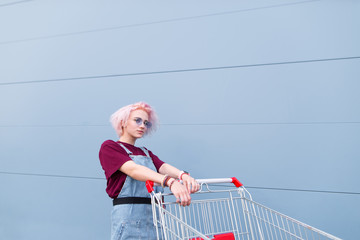 stylish bright girl with colored hair poses with a shopping cart on a blue background. Fashionable...