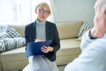 Adult woman in glasses taking notes on clipboard talking to elderly people in assisted living...