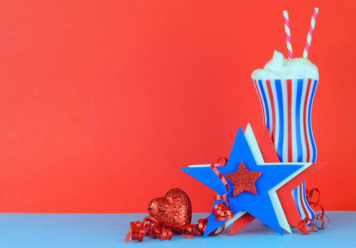Fun image for patriotic holidays in red white and blue. A striped glass with a whipped cream topped beverage and two straws on a red background and blue table top. Has star shapes and shiny ribbon.