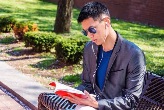 Young Asian American Man traveling, studying in New York, dressing in gray blazer, wearing white earphone, blue sunglasses, listening music, reading red book, sitting on bench in street park.. - Powered by Adobe