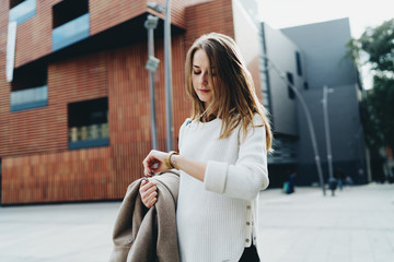 Young business woman looking on a watches while standing outdoors with a coat in her hand while waiting for a taxi. Student girl checking time while waiting for a friends outdoors.