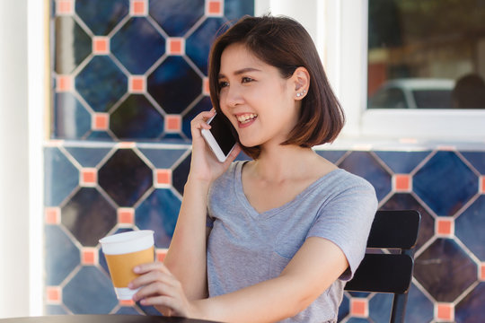Happy smile asian business women using talking cell phone sitting in cafe and holding a cup of coffee. Beautiful cheerful asian woman at cafe talking on phone and smiling while enjoying coffee.