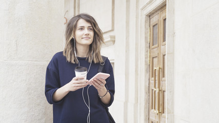 A smiling brunette white girl dressed in a blue sweatshirt is listening to music outdoors drinking a cup of coffee