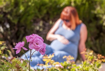 Happy young pregnant woman sitting in the park on a sunny day. Pregnancy expectation, love, care, rest, waiting for a baby concept. Blurry background, flower in the front.