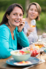 Looking at the camera. Portrait of a beautiful brunette woman in her forties. She sits around a table in a garden with friends for dinner.