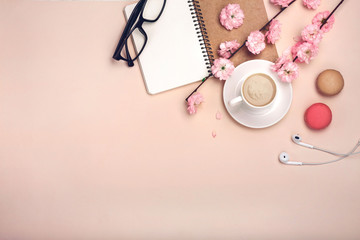 White cup with cappuccino, sakura flowers, macarons, notebook on a pastel pink background.