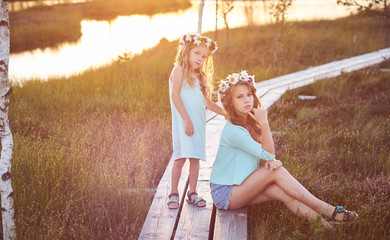 Two beautiful sisters standing against the background of a beautiful landscape, walk on the field near a pond at sunset.