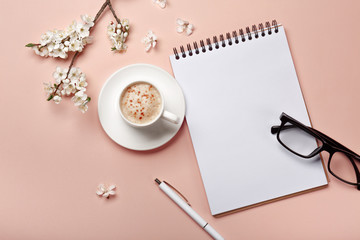 A white cappuccino cup with sakura flowers, notebook, headphones, pen and glasses on a pink background