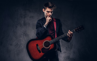 Handsome young thoughtful musician with stylish hair in elegant clothes posing with a guitar in his...