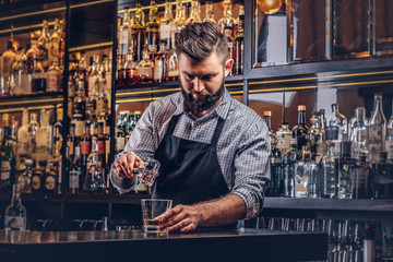 Stijlvolle brute barman in een shirt en schort maakt een cocktail op de achtergrond van de toog.