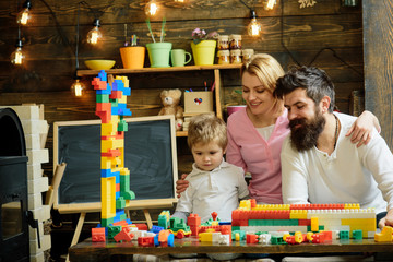 Concentrated boy building wall of colorful plastic blocks. Smiling mom and dad looking at adorable kid playing with construction bricks. Beautiful woman holding her son and husband by shoulders