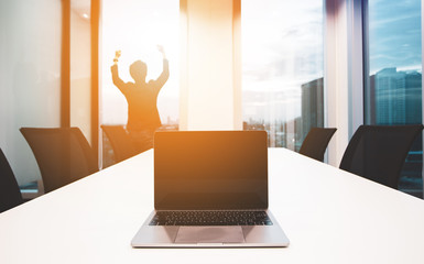 successful concept. modern laptop on foreground at meeting room with large windows. young business man are raising hands on background, outside building, city, tower view, soft focus