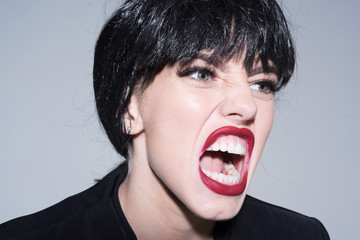 Woman with attractive red lips shouting, close up. Angry boss concept. Girl on scandalous shouting face wears formal jacket. Lady in black wig with make up screaming on grey background.