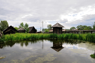 Obraz premium A small swampy pond in the village. Old wooden houses, bulrush. Cloudy summer day.