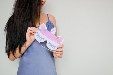 Brunette girl in a blue nightdress holds a mask for sleeping with a unicorn in her hands. Copy space