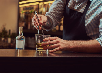 Stylish brutal barman in a shirt and apron makes a cocktail at bar counter background.
