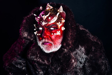 Senior man with white beard dressed like monster. Alien, demon, sorcerer makeup. Evil concept. Demon with red face on black background, close up. Man with thorns or warts in fur coat.