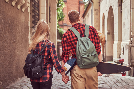 Back view of a young hipster couple, handsome skater and his girlfriend, holding hands, walking around a old narrow streets of Europe.
