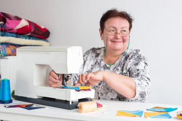 manufactoring, handcraft, profession concept. there is an old smiling woman wearing glasses and white shirt with grey floral patterns, she is sitting at her work desk by electronic sewing machine