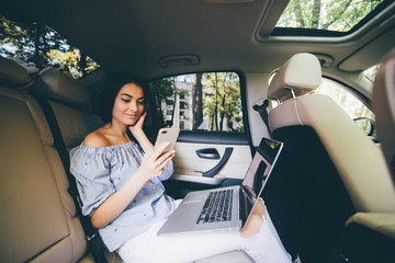 Attractive young business woman working on her laptop at the back sit of a car and talking on the phone on the way to meeting