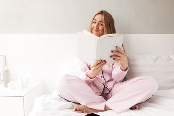 Portrait of young smiling woman sitting on bed and joyfully looking in book at home isolated