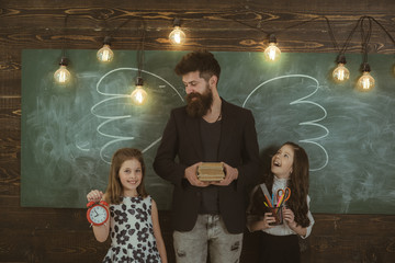 Favourite teacher concept. Teacher and girls pupils in classroom, chalkboard on background. Children and teacher with drawn by chalk wings. Man with beard and schoolgirls with school attributes.
