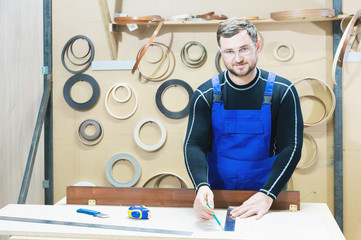 A bearded tired handsome carpenter on a table with a pencil draws a sign on the board. At the...
