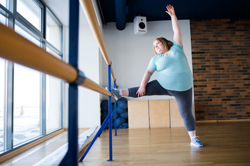 Full length portrait of obese young woman doing ballet poses stretching at gymnastic bar in dance class, copy space